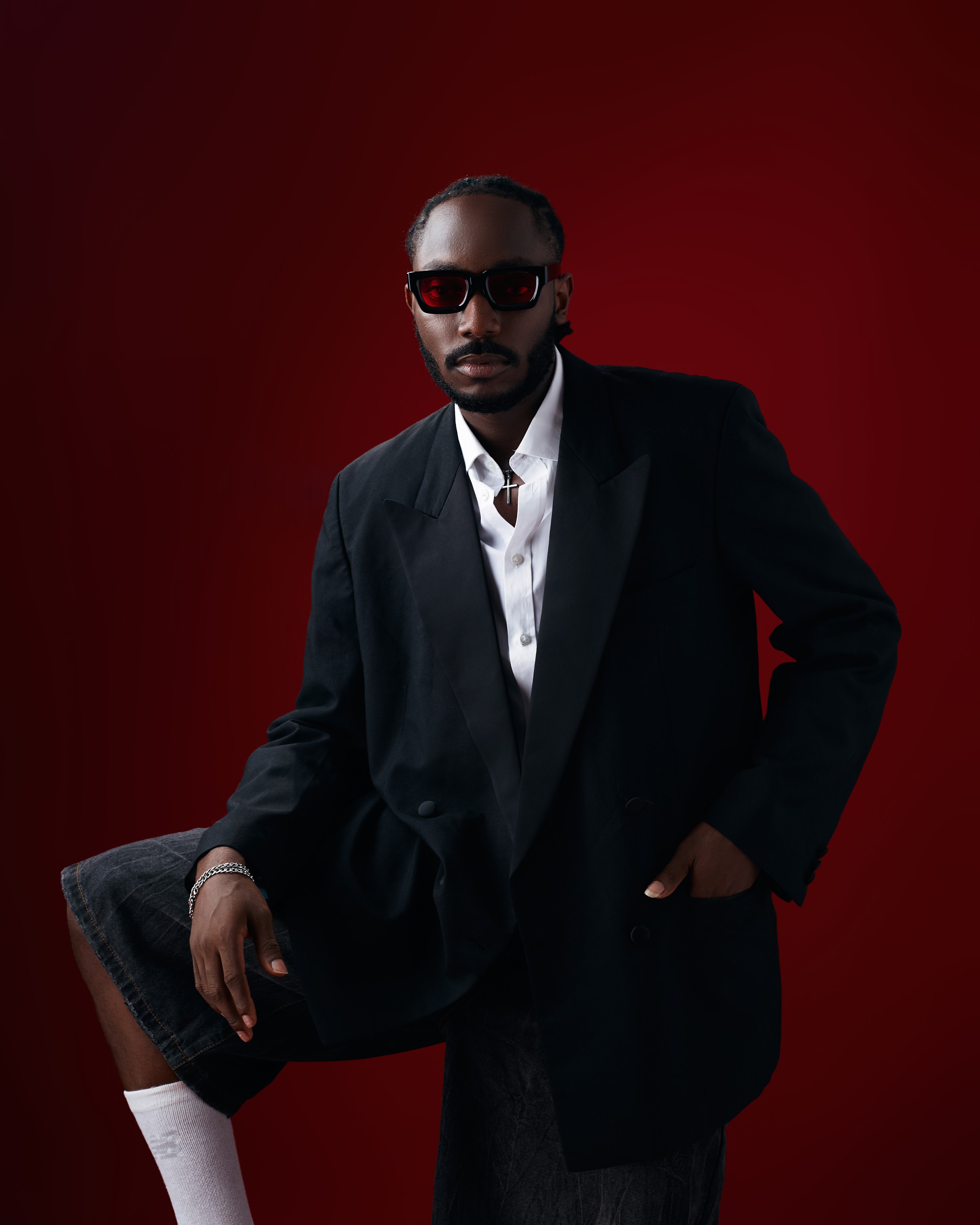 Saelem in a black suit and red-tinted sunglasses against a deep red studio backdrop